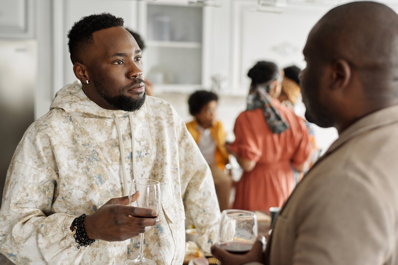 Two friends having a discussion at a casual indoor gathering with others in the background.